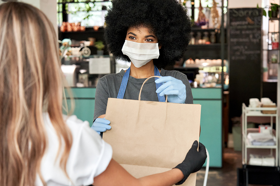 Black woman wearing surgical mask and gloves with beautiful 4C hair hands takeout bag to white woman customer in gloves