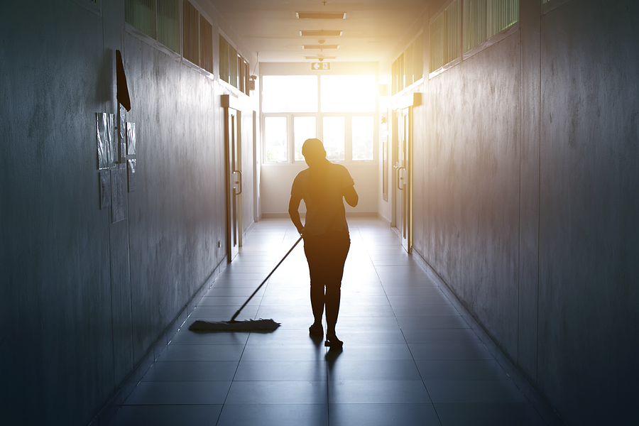 Custodian pushes a push broom in backlit hallway