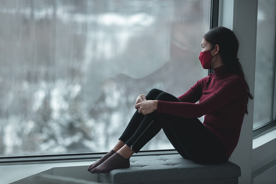 Woman in COVID mask sits at window alone, looking out at a gray city scene