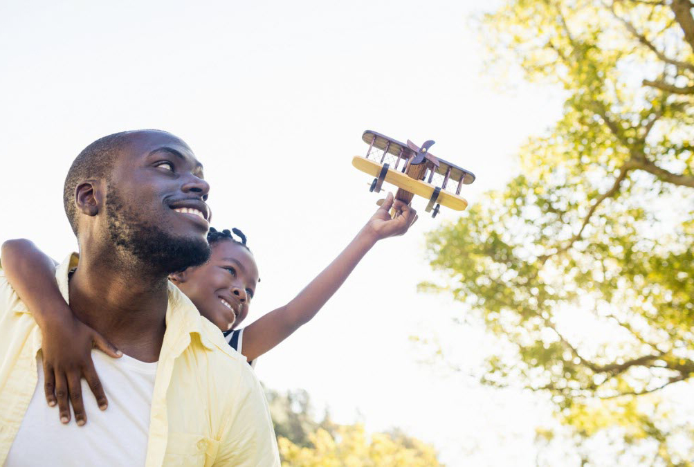 African-American father holds child flying toy airplane in bright sky
