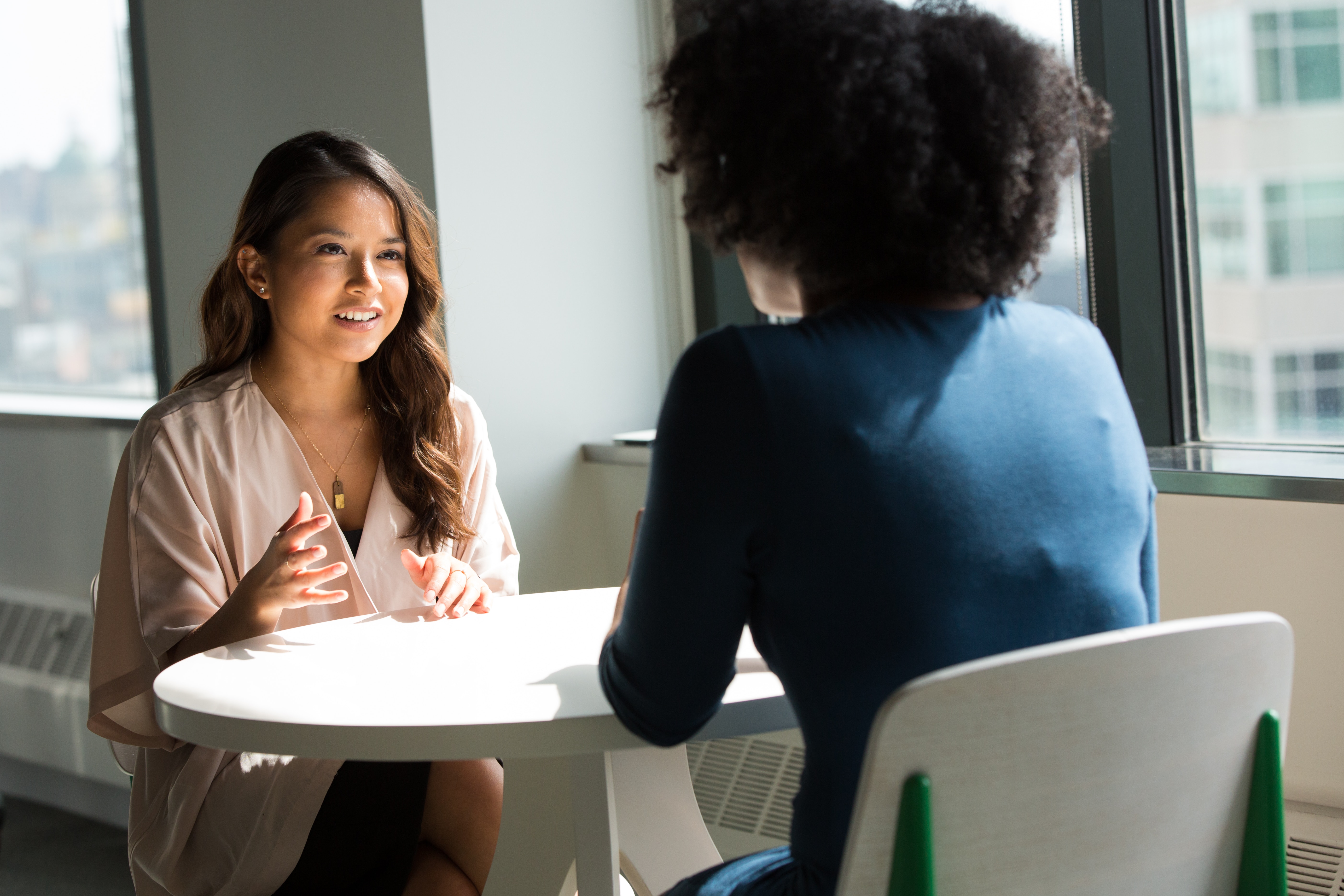 Two women talk at table, one seen in foreground from behind. At window, office buildings beyond. 