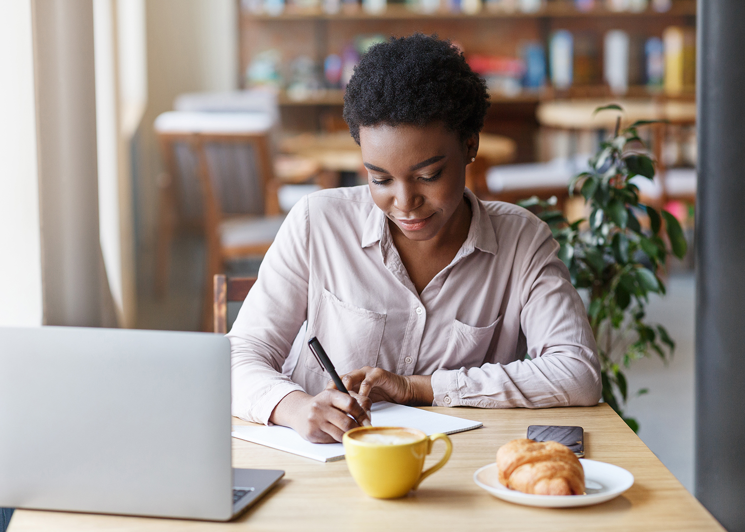 Woman works on her dissertation in a cafe with a debranded Macbook, a 12-ounce caffe latte and a large croissant. She writes on a pad of paper, looking pleased with her train of thought. 