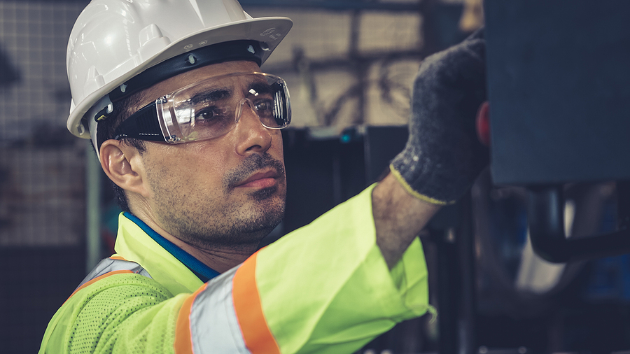 Factory worker in high-vis jacket and protective helmet and glasses pushes button with gloved hand, looking content but serious