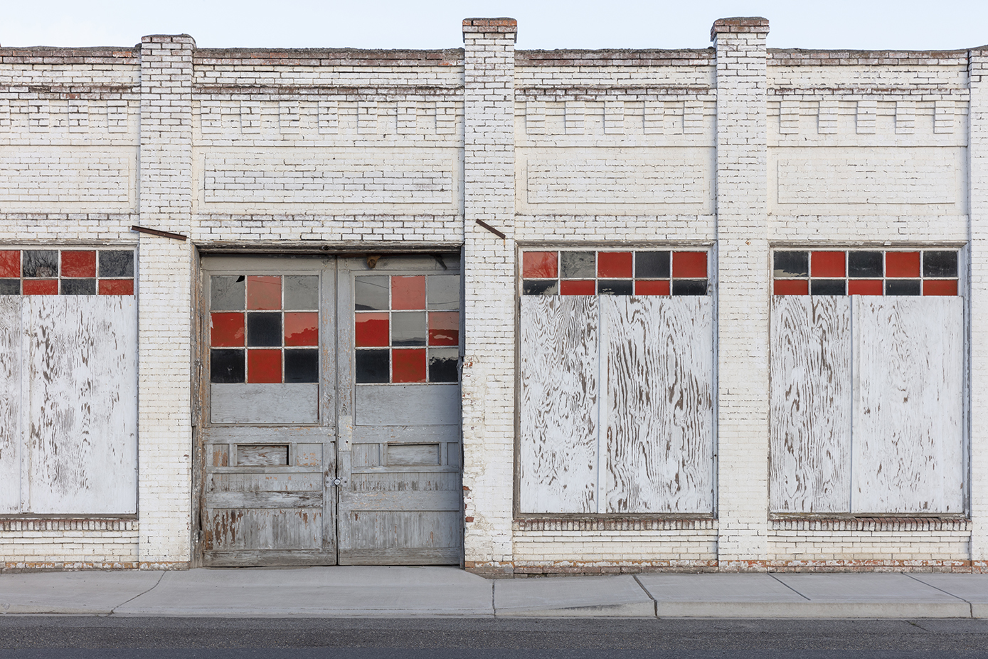 White brick building with boarded windows, also whitewashed, looks like on a Main street. Some decorative checkerboard pattern visible on windows above boards