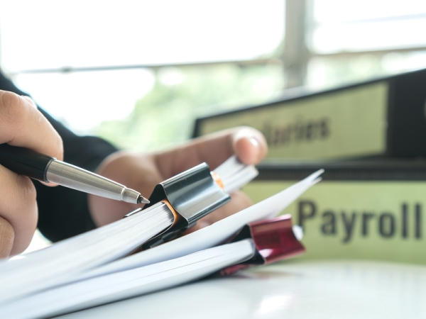 Hands, pen, paper with binder clips, binder reading "payroll"
